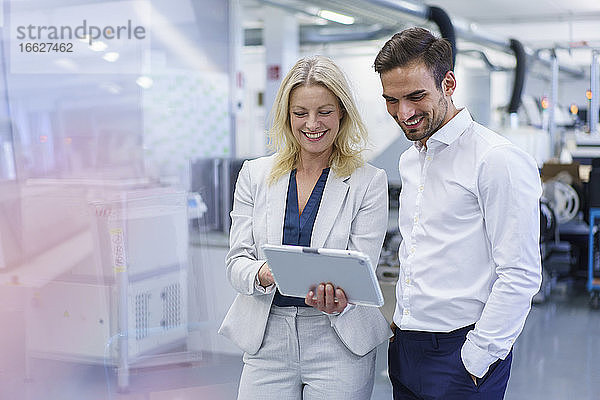 Lächelnde blonde Geschäftsfrau  die mit einem jungen männlichen Kollegen in einer Fabrik über ein digitales Tablet diskutiert