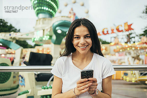 Nahaufnahme einer lächelnden jungen Frau  die ihr Smartphone vor einer Fahrt im Vergnügungspark benutzt