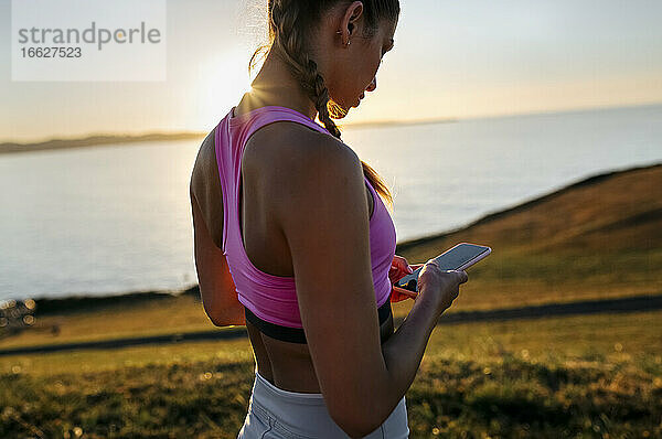 Junge Frau benutzt ihr Smartphone  während sie auf einem Hügel bei Sonnenuntergang am Meer steht