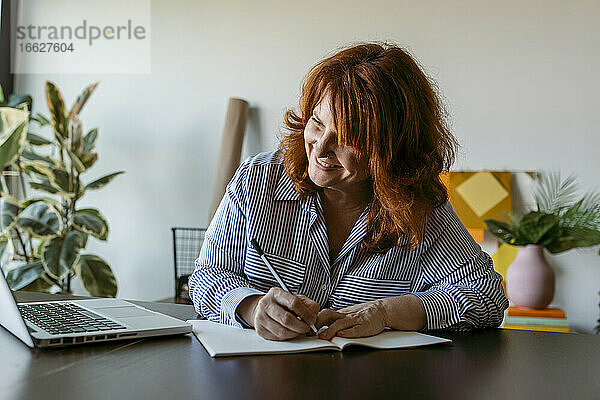 Lächelnde Frau schreibt ein Buch  während sie zu Hause am Tisch sitzt