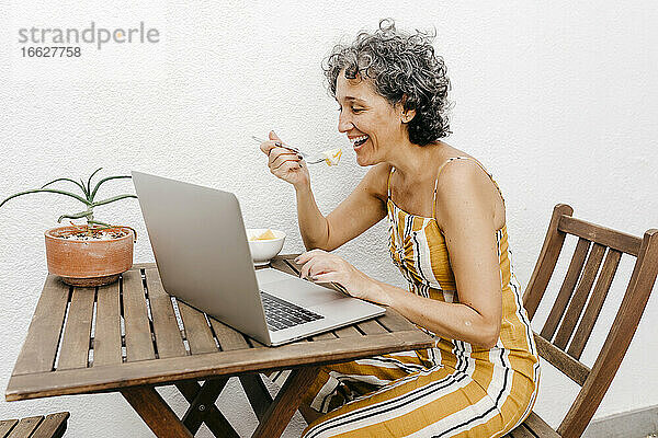 Lächelnde reife Frau  die an einem Tisch an einer weißen Wand im Hinterhof isst und dabei einen Laptop benutzt