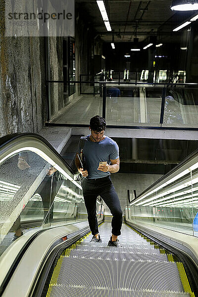 Männlicher Pendler  der ein Mobiltelefon benutzt  während er auf einer Rolltreppe in der U-Bahn steht