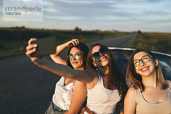 Lächelnde Freunde  die ein Selfie machen  während sie bei Sonnenuntergang auf der Straße stehen