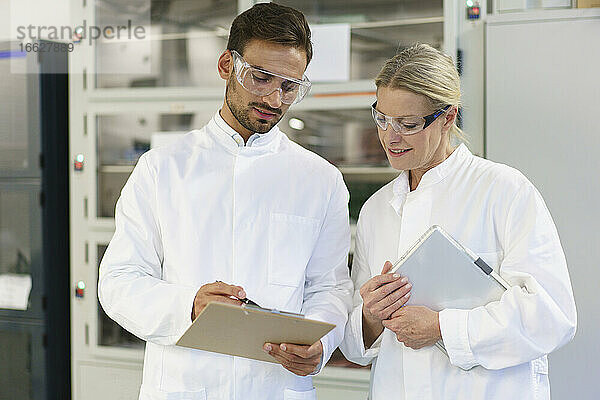 Junger männlicher Techniker  der mit einer blonden Kollegin  die ein digitales Tablet hält  in einem Labor über ein Klemmbrett diskutiert