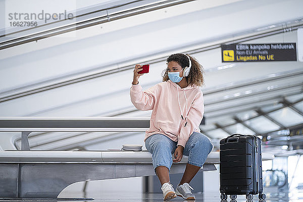 Junge Frau  die ein Selfie macht  während sie über Kopfhörer am Flughafen Musik hört