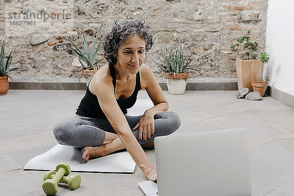 Lächelnde Frau mit Laptop für das Lernen Yoga durch Online-Tutorial  während im Hinterhof sitzen