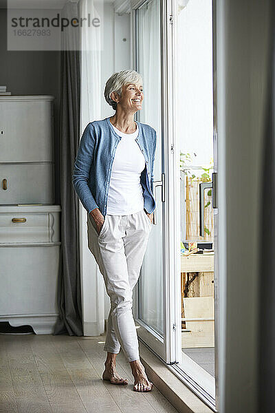 Ältere Frau steht mit den Händen in den Taschen und schaut aus dem Fenster zu Hause