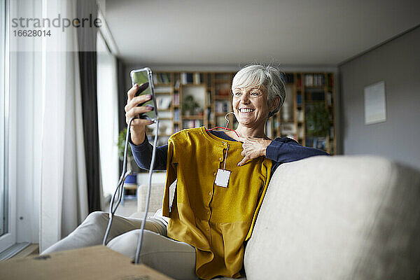 Lächelnde ältere Frau  die ein Selfie mit neuer Bluse auf dem Smartphone macht  während sie zu Hause sitzt