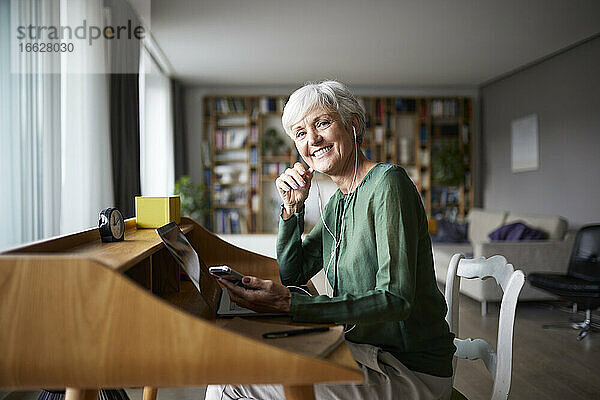Lächelnde aktive ältere Frau hört Musik  während sie zu Hause auf einem Stuhl sitzt