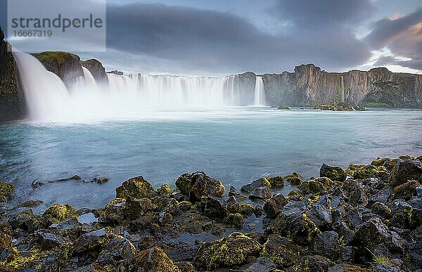 Langzeitbelichtung eines türkisen Wasserfalls in vulkanischer Landschaft mit dramatischen Wolken und grünem Moos auf Felsen  Godafoss  Þingeyjarsveit  Norðurland eystra  Island  Europa