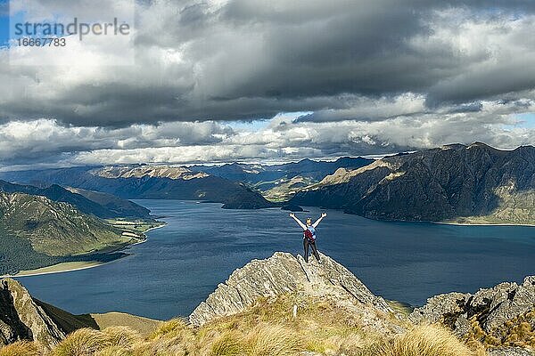 Wanderin steht auf einem Felsen und streckt die Arme in die Luft  Ausblick über Lake Hawea  See und Berglandschaft im Abendlicht  Ausblick vom Isthmus Peak  Wanaka  Otago  Südinsel  Neuseeland  Ozeanien