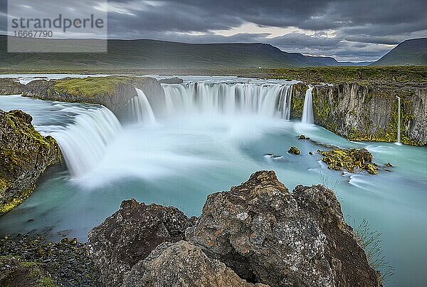 Langzeitbelichtung eines türkisen Wasserfalls in vulkanischer Landschaft mit dramatischen Wolken  Godafoss  Þingeyjarsveit  Norðurland eystra  Island  Europa