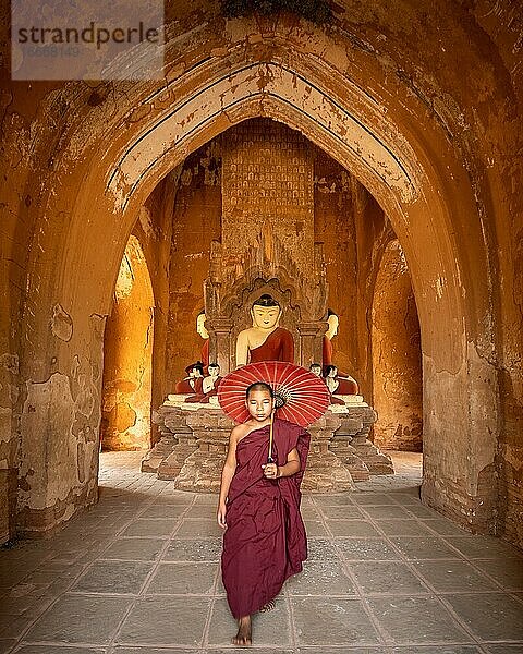 Buddhistischer junger Mönch in roter Robe mit rotem Schirm geht in einem Tempel  Bagan  Myanmar  Asien