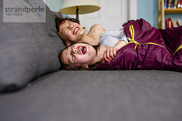zwei glückliche Kinder liegen zusammen auf der Couch und lachen lauthals vor Freude