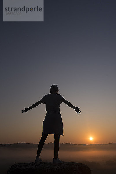 Silhouette eines Mädchens auf einem Berggipfel mit ausgestreckten Armen bei Sonnenaufgang