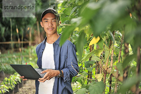 Junger asiatischer Bauer mit Laptop im Garten