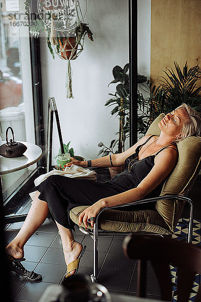 Blonde Frau in schwarzem Kleid sitzt allein im Café in entspannter Haltung