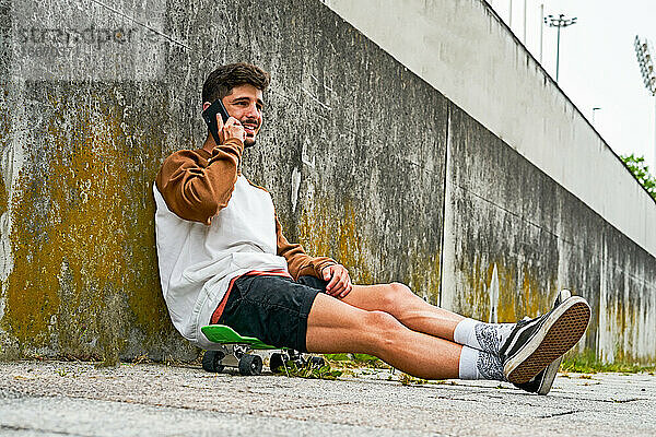 Junger lässiger Mann sitzt im Freien auf einem Skateboard und benutzt ein Telefon