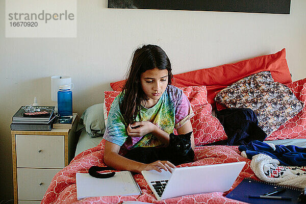 Mädchen mit ihrer schwarzen Katze macht Schularbeiten im Bett auf ihrem Laptop
