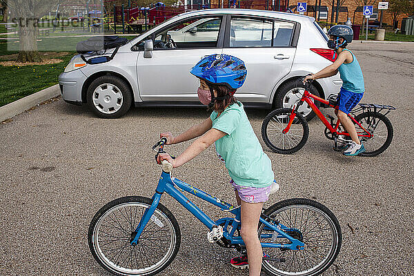 Ein Junge und ein Mädchen mit Gesichtsmasken fahren zusammen auf einem Parkplatz Fahrrad