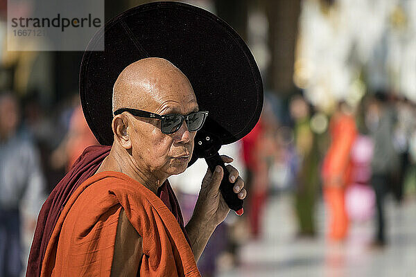 Buddhistischer Mönch in safranfarbener Robe mit Sonnenbrille und Handfächer an einem sonnigen Tag  Shwedagon-Pagodenkomplex  Yangon  Myanmar