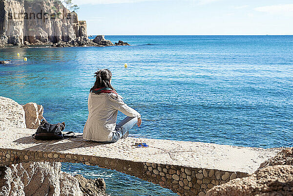 Rückansicht einer Frau  die sich vor dem blauen Meer entspannt  auf einer Brücke sitzend