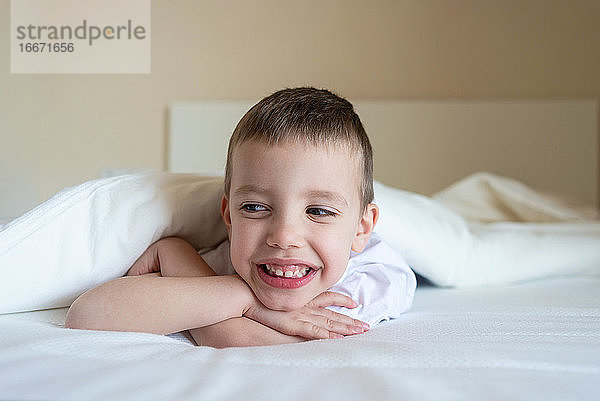 Bezauberndes lächelndes Kind  das unter einer Decke auf dem Bett liegt und in die Kamera schaut