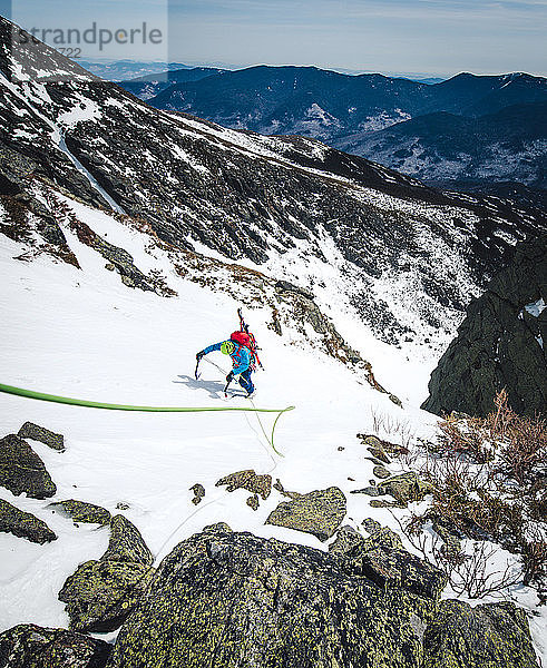 Mann klettert mit Kletterseil und Skiern eine steile Schneerinne hinauf