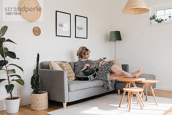 Frau saß in einem Kleid auf der Couch und spielte zu Hause Gitarre und entspannte sich