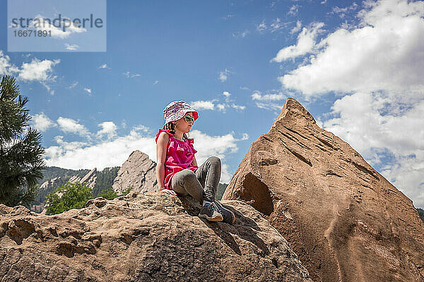 junges Mädchen mit Hut sitzt auf einem Felsen mit Bergen im Hintergrund nach einer Wanderung