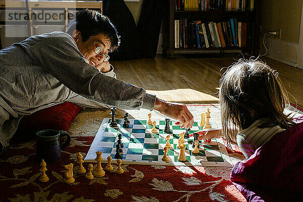 Ein lächelnder Vater liegt auf dem Boden mit einem kleinen Mädchen  das im Sonnenschein Schach spielt