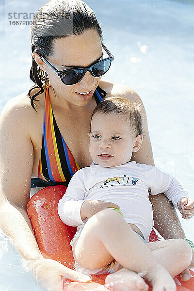 Mutter trägt Sonnenbrille und schwimmt mit ihrem kleinen Sohn in einem Pool an einem sonnigen Tag