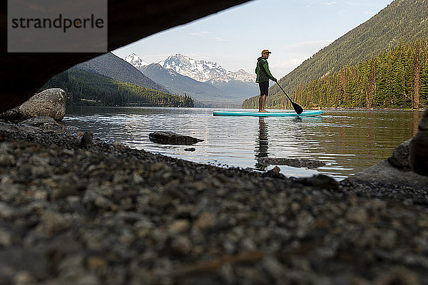 Ebenerdige Seitenansicht eines männlichen Reisenden  der auf einem SUP-Board auf dem friedlichen Duffey Lake vor dem Hintergrund der Berge am Morgen in British Columbia steht