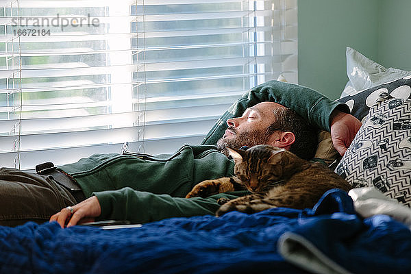Mann macht ein Katzennickerchen mit seiner getigerten Katze  die neben ihm kuschelt