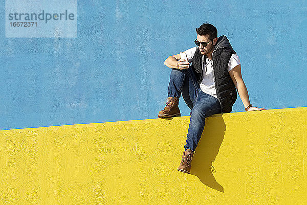 Junger bärtiger Mann mit Sonnenbrille sitzt an einer gelben Wand und benutzt ein Telefon