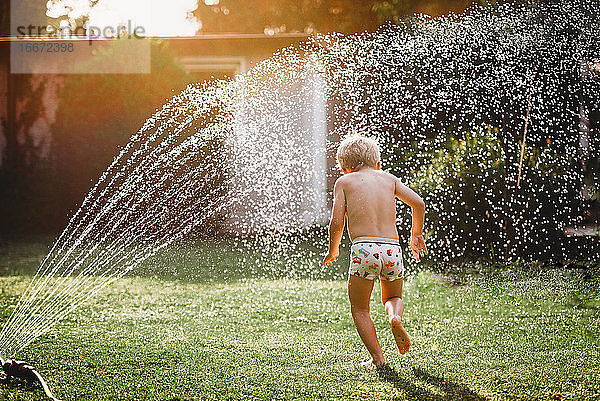 Junge weiße Junge läuft unter dem Wasser aus dem Sprinkler im Garten