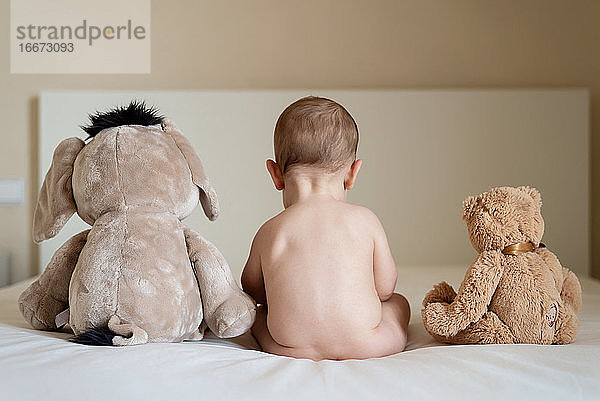 Rückansicht eines nackten Babys  das mit zwei Teddybären im Bett liegt