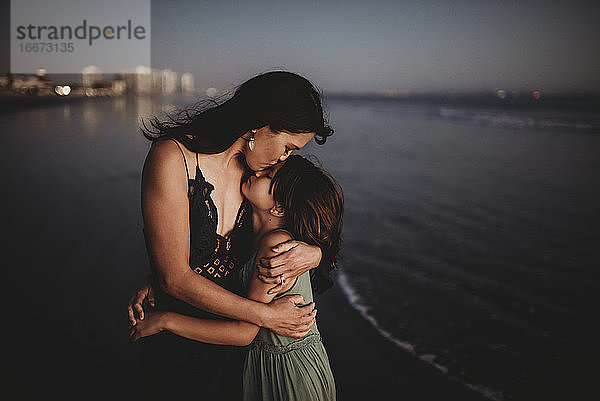 Zärtliche Mutter umarmt und küsst 8 Jahre alte Tochter am Strand bei Sonnenuntergang