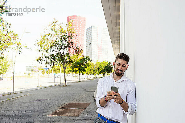 Junger bärtiger Mann lehnt an einer weißen Wand und benutzt ein Mobiltelefon