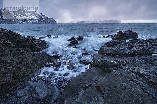 Die schroffen Berge von Flakstadøy erheben sich vor der felsigen Küste von Vareid  Lofoten  Norwegen  über das Meer.