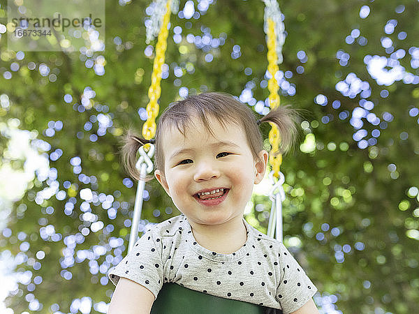 Happy Smiling Kleinkind Mädchen mit Zöpfen Swings im Freien unter einem Baum