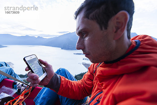 Perplexer Mann  der bei Sonnenuntergang auf dem Portaledge sitzt und auf sein Telefon schaut