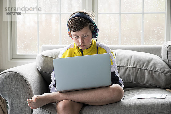 Junge mit Laptop für virtuelle Schule sitzt auf der Couch im Wohnzimmer