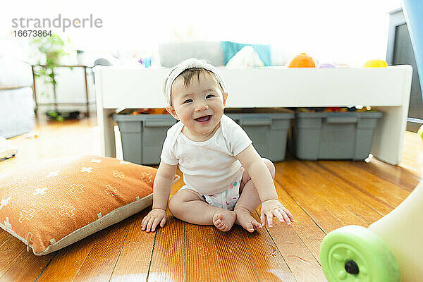 Glücklich lächelndes Kleinkindmädchen sitzt zu Hause auf dem Boden  umgeben von Spielzeug