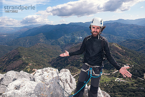 Konzept: Abenteuer. Bergsteiger Mann mit Helm und Gurtzeug. Auf dem Gipfel des Berges. Mit offenen Armen und lächelnd zeigt er die Landschaft. Via ferrata auf Felsen.