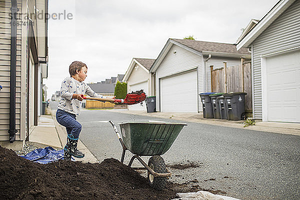 Kleiner Junge hebt eine Schaufel Erde in eine Schubkarre in einer Seitengasse
