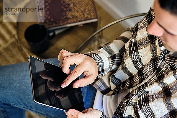Junger Mann  der zu Hause sitzt und ein digitales Tablet benutzt. Chillt am Fenster und konsumiert digitale Inhalte. Ansicht von oben.