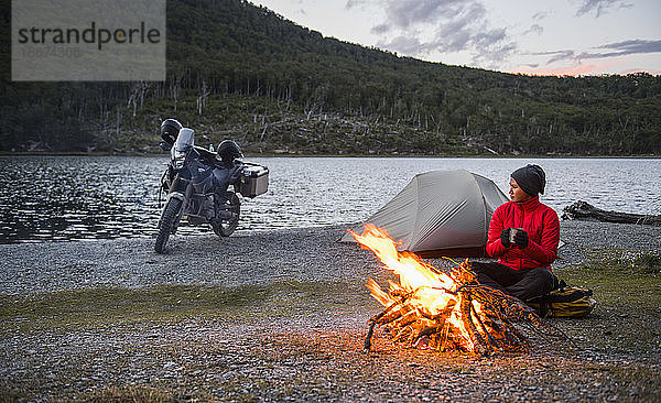 Frau genießt das Lagerfeuer im Camp neben einem stillen See in Feuerland