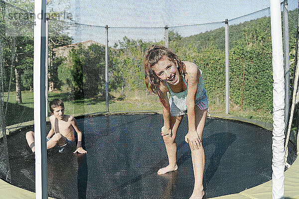 Horizontales Foto eines blonden Jungen und eines brünetten Mädchens  die in die Kamera schauen und sich auf einem Trampolin vor einem grünen Hintergrund vergnügen. Der Teenager und das Kind tragen kurze Jeans und sie sehen glücklich aus
