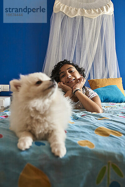 ein kleines Mädchen und ihr weißer Hund im Vordergrund  die auf dem Bett sitzen und in die Kamera schauen